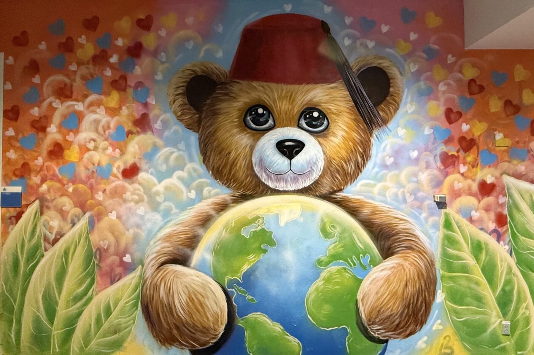 An airbrushed mural of a teddy bear in a maroon Shriners fez hugging a glowing globe, on a sunset-orange wall scattered with hearts.